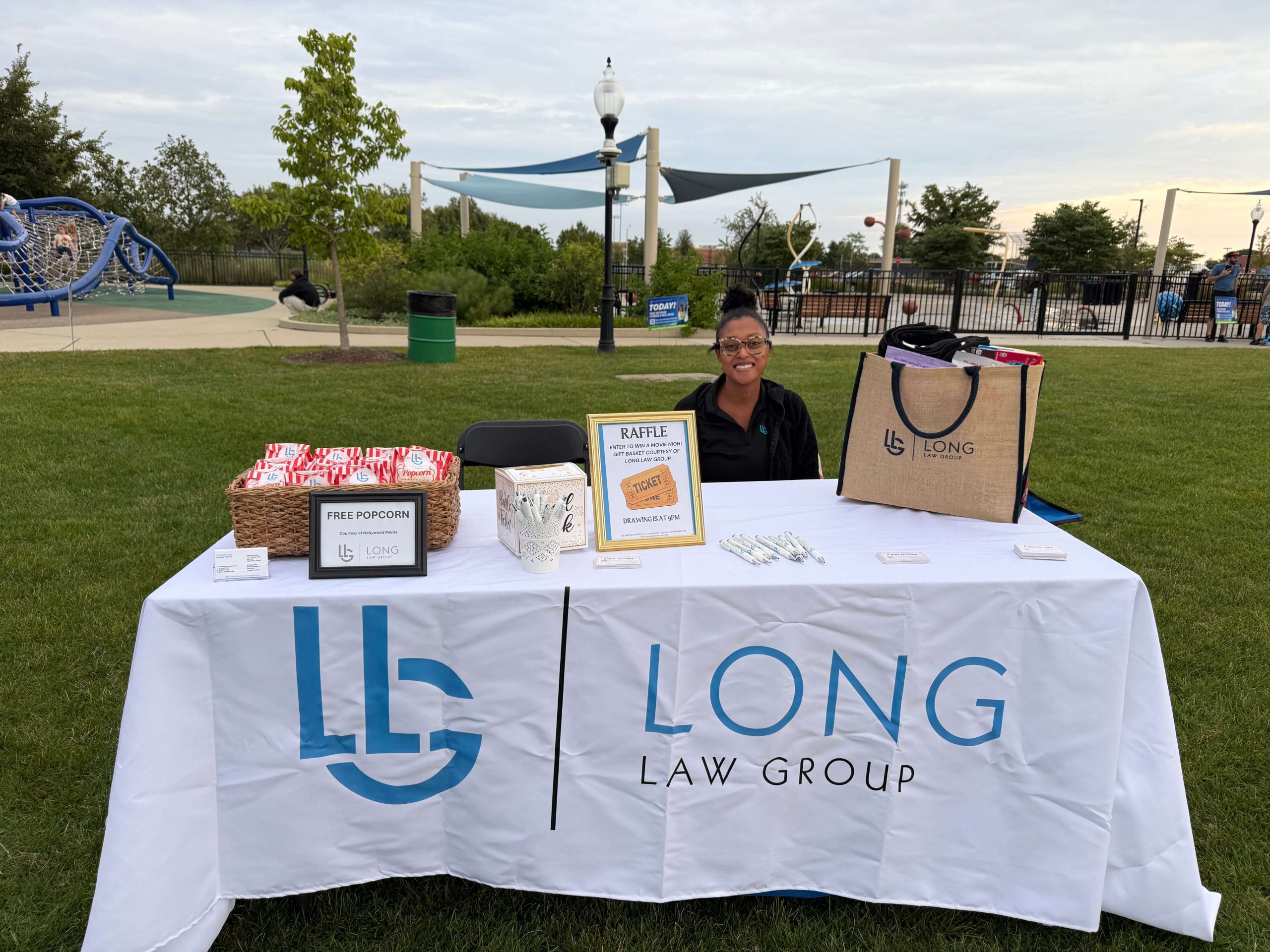 Long Law Group team member at community event booth with branded table, free popcorn basket, raffle prizes and park playground in background